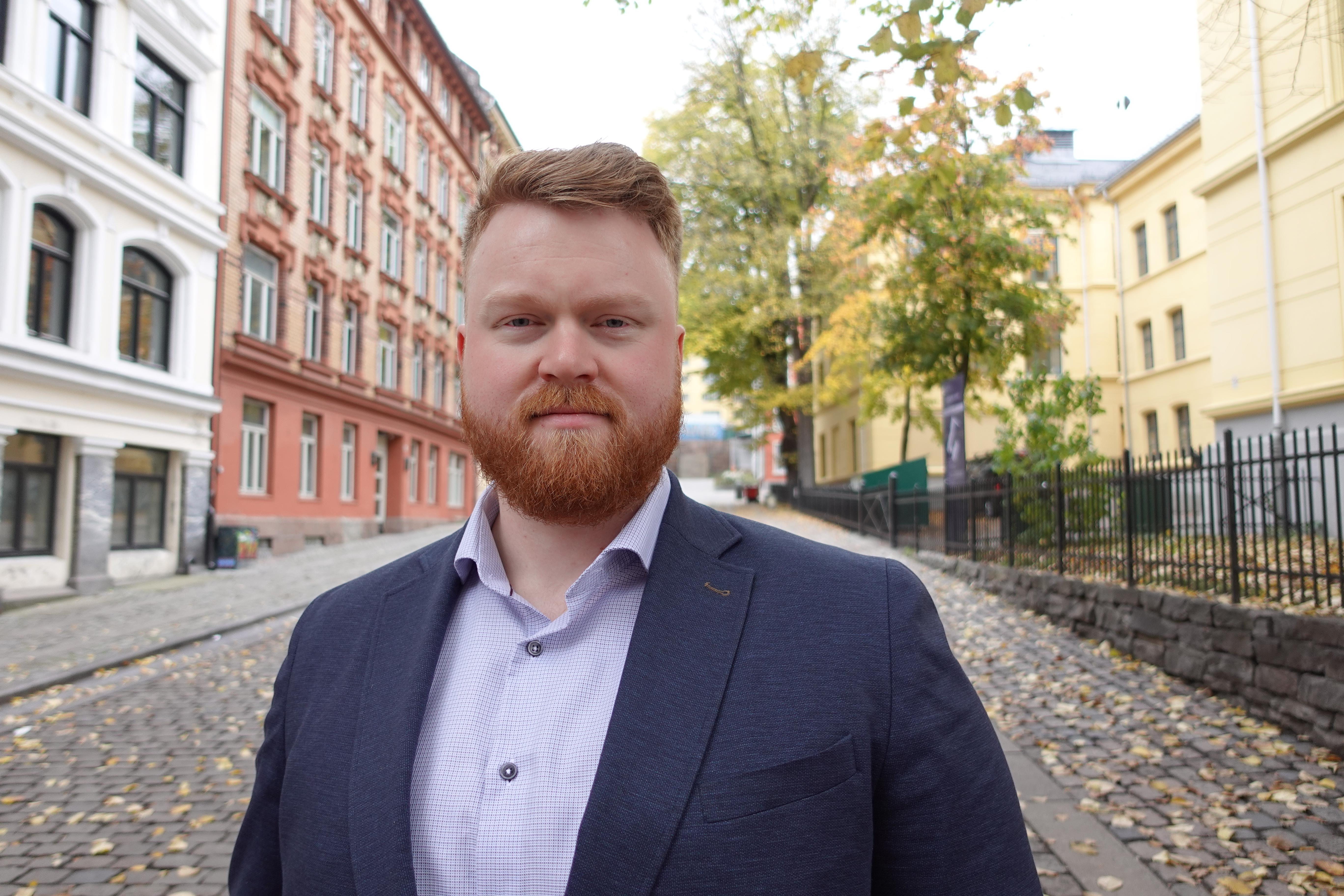 Bilde av Lars Strande Syrrist. Smilende mann med rødt hår og skjegg, kledd i lys skjorte og mørk blazer. Står foran gate med flere bygg. 