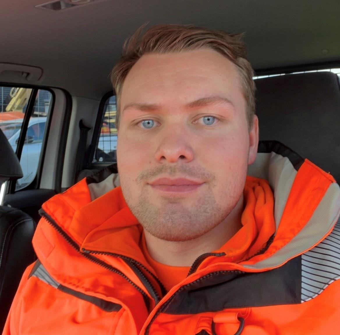 Bilde av Kasper Faugli Åvitsland. Mann som smiler mot kamera, har lyst hår og blå øyne. Han har på seg en oransje jakke og sitter inne i en bil. 