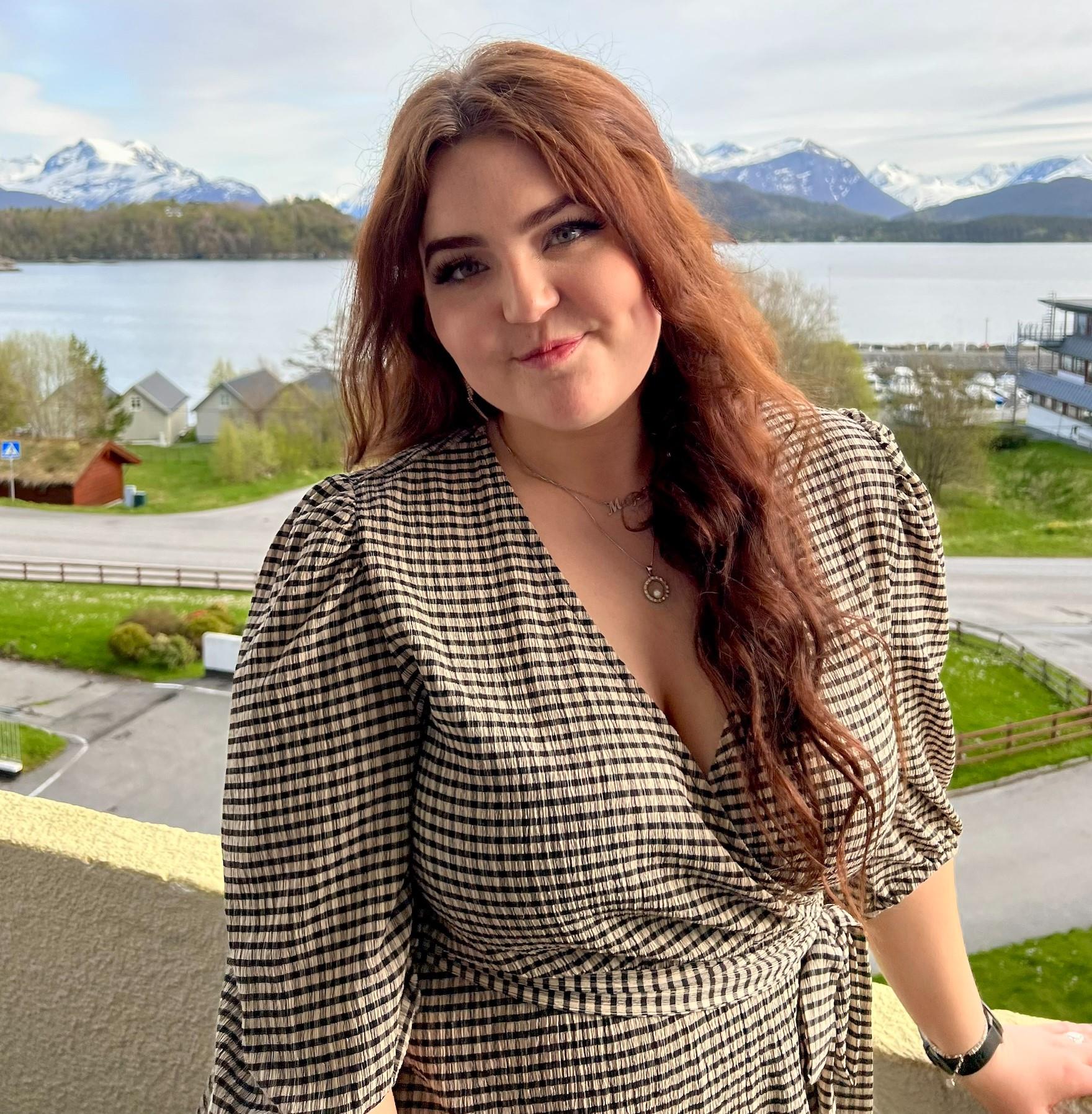 Bilde av Malin Svarstad. Kvinne som smiler mot kamera. Hun har rødt hår og har på seg en rutete kjole. Hun står ute på en terrasse, og man ser fjell og hav i bakgrunn. 
