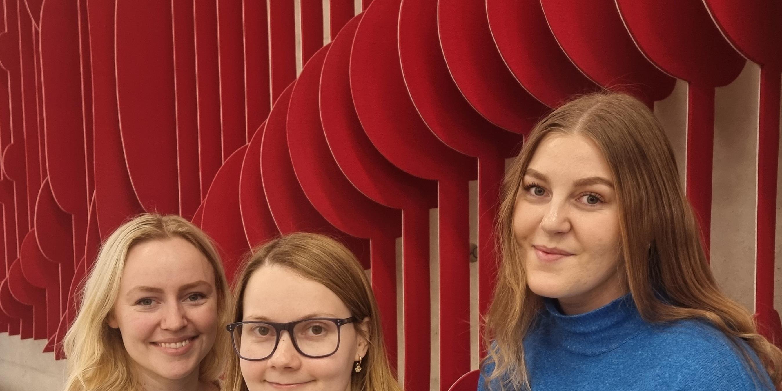 Tre kvinner smiler til kameraet. Bak dem er en rød utsmykning på veggen. Foto.