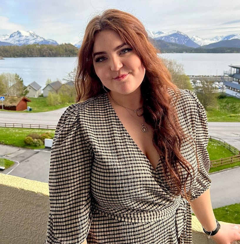Bilde av Malin Svarstad. Kvinne som smiler mot kamera. Hun har rødt hår og har på seg en rutete kjole. Hun står ute på en terrasse, og man ser fjell og hav i bakgrunn. 